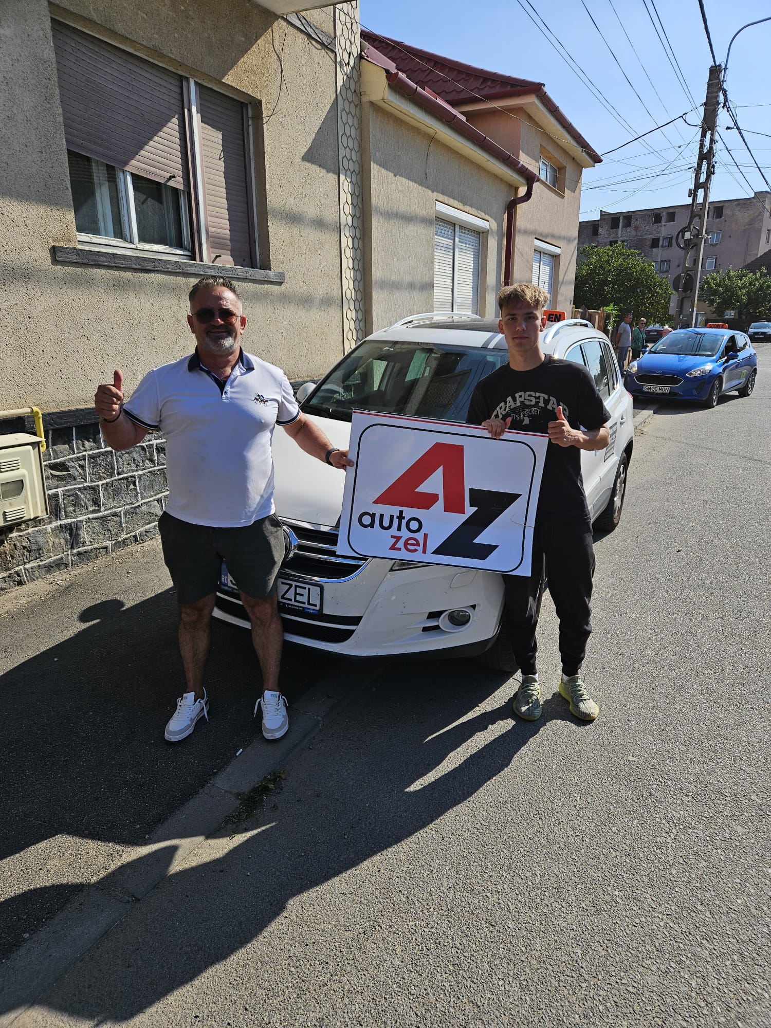 Instructor Auto Zel și cursant cu degetele mari ridicate lângă mașina școlii